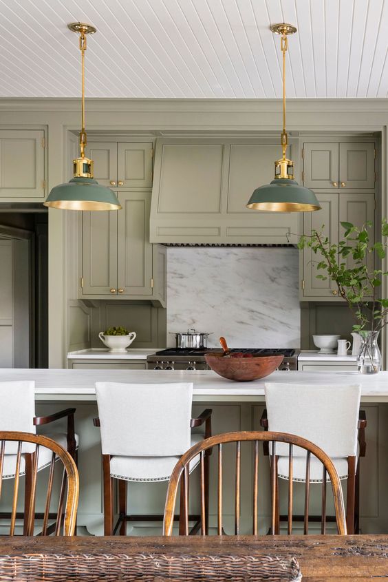 a pretty green kitchen with shaker cabinets, a white stone backsplash and countertops, white stools, green and brass pendant lamps and brass fixtures