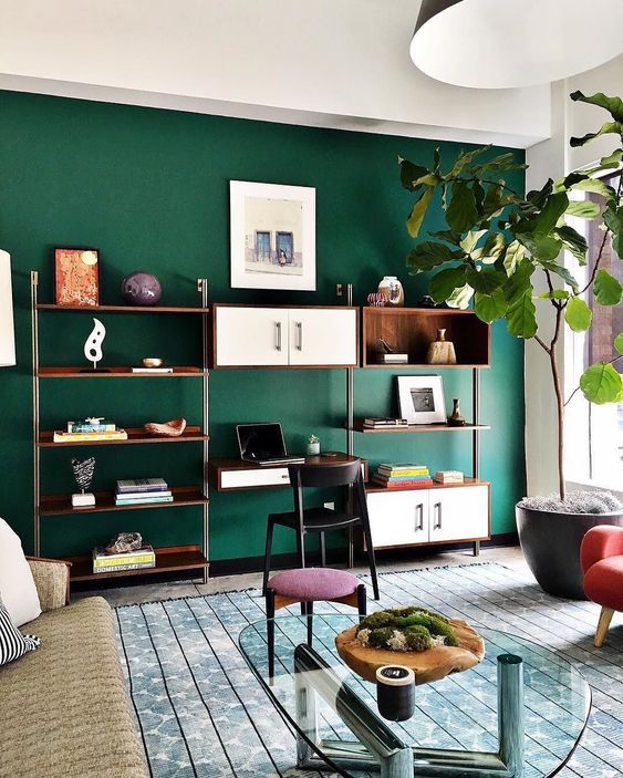 a pretty living room with an emerald accent wall, neutral sofa, jewel tone seating furniture, an open shelving unit and a potted tree