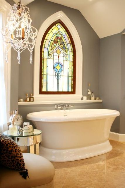 a refined bathroom with grey walls, a raised oval tub, a chic stained glass window in Gothic style and a crystal chandelier is wow