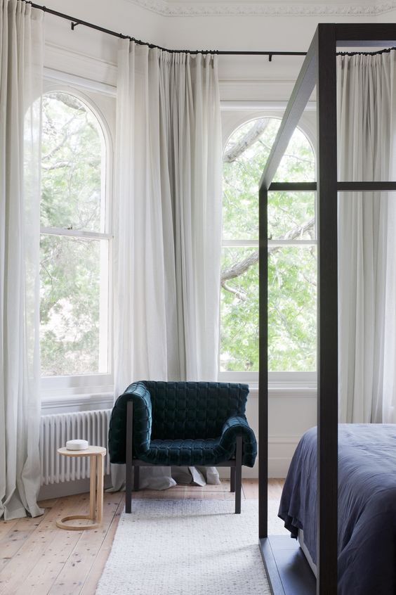 a refined contemporary bedroom done with arched windows, a black frame bed, a navy chair and some neutral and flowy curtains