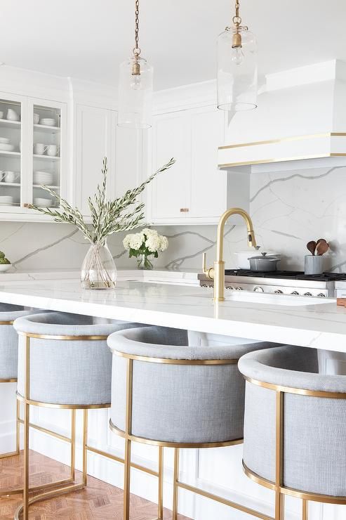 a refined neutral kitchen with white cabinets, a white marble backsplash and countertops, brass fixtures and tall stools with brass framing