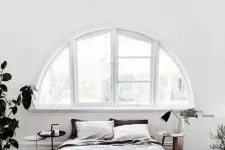 a relaxed Scandinavian bedroom with a white frame arched window, a bed and a bench, some nightstands and a statement potted plant