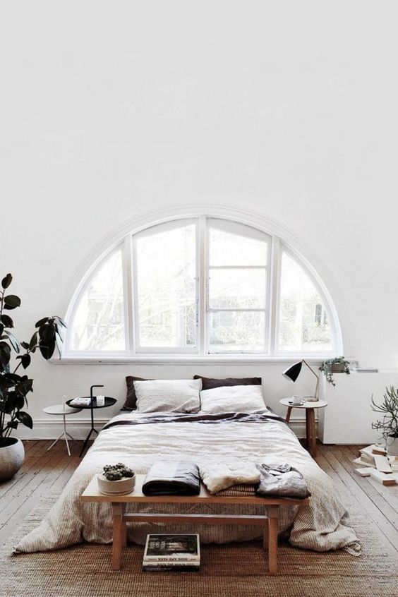 a relaxed Scandinavian bedroom with a white frame arched window, a bed and a bench, some nightstands and a statement potted plant