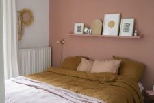 a relaxed and welcoming bedroom with a dusty pink accent wall, a bed, a long open shelf, a nightstand, a woven pendant lamp, some art and mustard and pink bedding