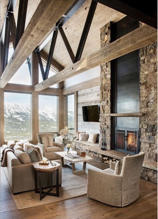 a rustic cabin living room with wooden beams, a fireplace clad with stone, tan seating furniture, a coffee table and a side one that matches