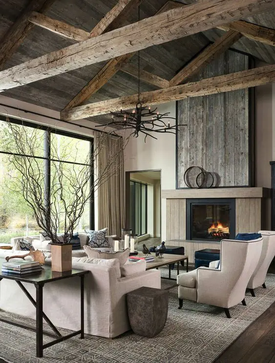 a rustic living room with weathered wooden beams and a ceiling, a fireplace clad with the same wood, neutral seating furniture, black poufs and a console table