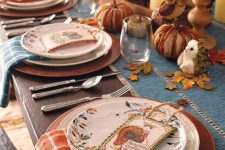 a rustic vintage Thanksgiving tablescape with a blue table runner and blue and orange plaid napkins, faux pumpkins, pillar candles and floral plates