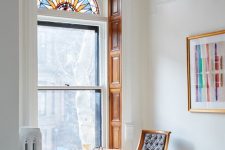 a small and lovely nook with a tall window with stained glass in the upper part, a coffee table and a chair and a bold artwork that echoes with the window