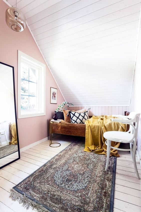 a small attic bedroom with a pink accent wall, a bed with colorful bedding, a white chair, a mirror in a black frame and a boho printed rug