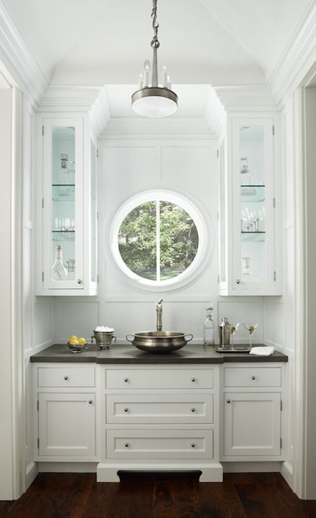 a small home bar styled with traditional white cabinets and a small porthole window to make the space unique