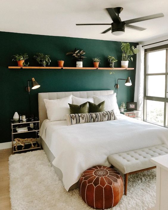 a stylish boho bedroom with a dark green accent wall, a long shelf with potted plants, a neutral upholstered bed and a bench, a brown Moroccan pouf and glass nightstands