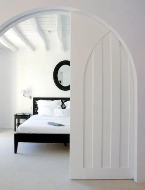 a totally white space with a black bed, mirror and nightstands and an arched pocket door that looks both modern and antique at the same time