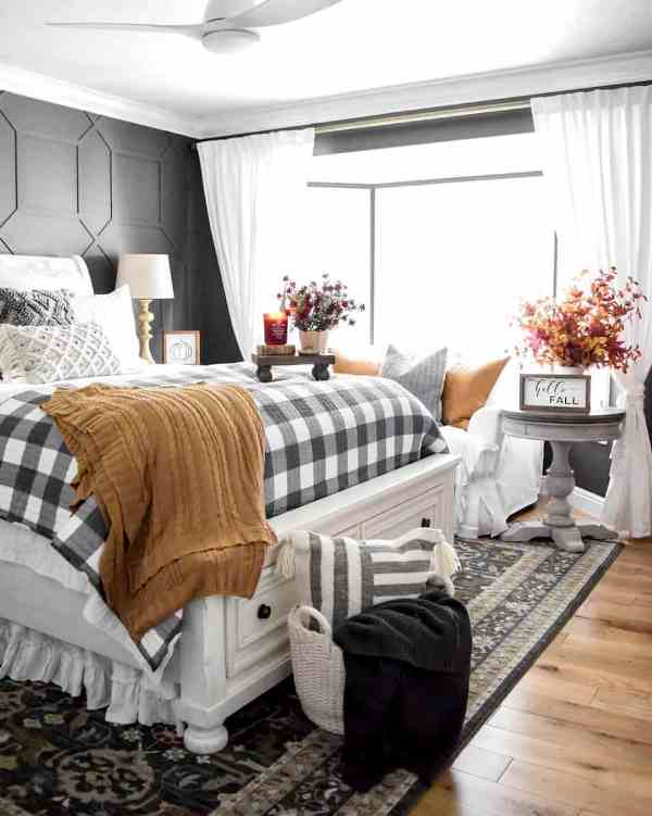a vintage famrhouse bedroom with black paneled walls, a white bed with drawers, plaid and mustard bedding, a basket with a pillow and some fall leaf arrangements
