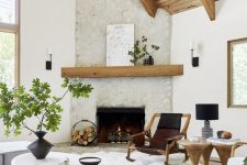 a welcoming bright living room with a fireplace clad with stone, wooden beams, catchy furniture, a large rug and a knit pouf plus greenery