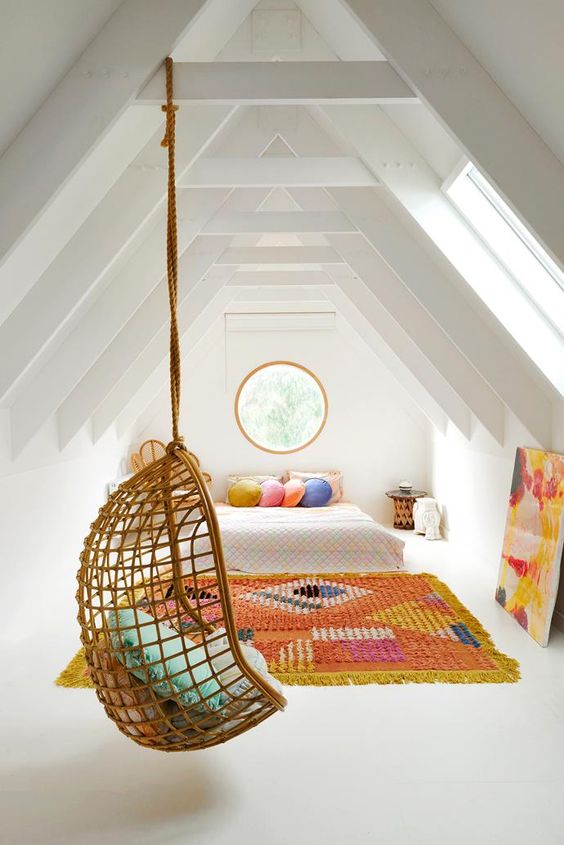 a white attic bedroom with bright textiles, a round window, bold pillows and a cool rattan pendant chair is amazing
