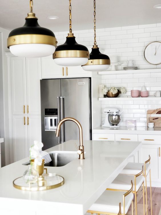 a white kitchen with shaker cabinets, a kitchen island with tall stools, black and brass pendant lamps, brass fixtures and stools with brass framing