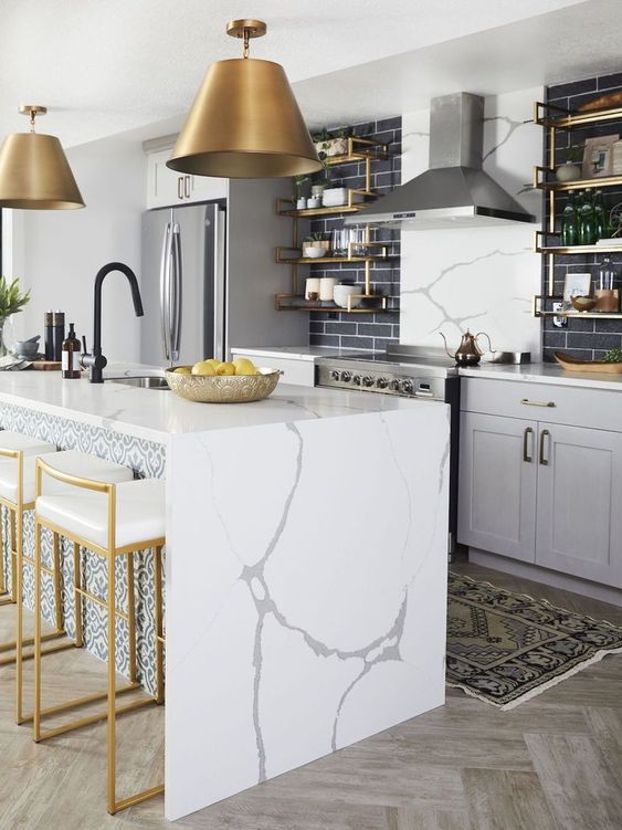 an elegant grey kitchen with shaker cabinets, black skinny tiles, a kitchen island with a waterfall countertop, brass lamps, stools and shelves