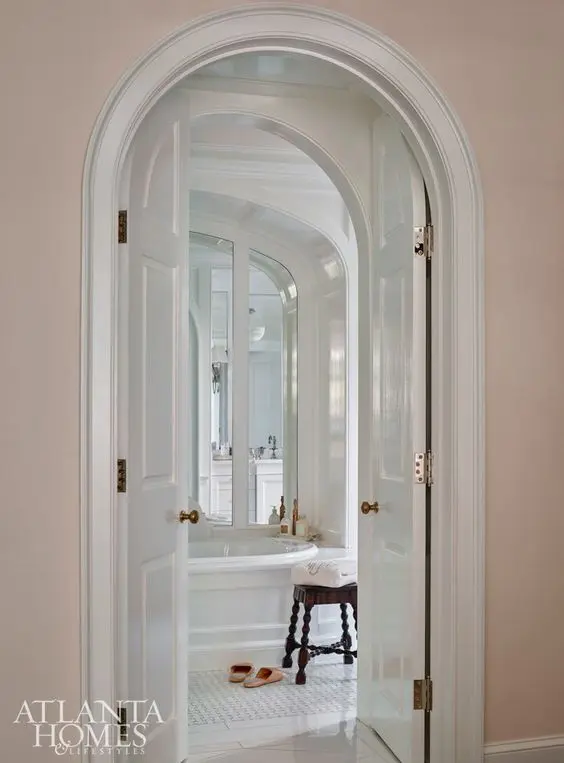 double-height arched doors leading to a refined white bathroom make it look super chic and super cool at the same time