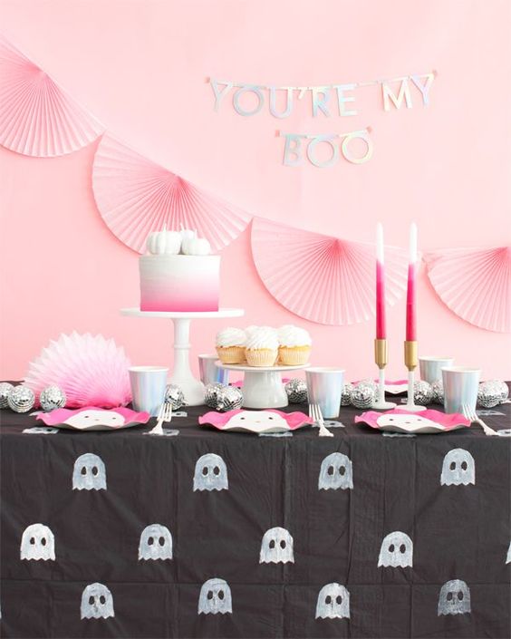 a pretty Halloween sweets table in pink and black, with pink paper fans, pink candles and a cake, pink ghost plates and a ghost tablecloth