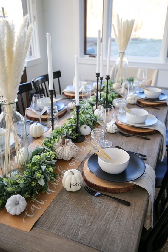 a beautiful rustic Thanksgiving table setting with a greenery runner, white faux pumpkins, black plates and white bowls, grasses in vases