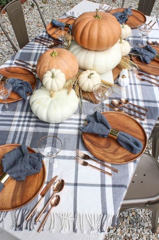 a beautiful rustic Thanksgiving tablescape with a plaid tablecloth, wooden chargers, stacked pumpkins, wheat and blue napkins