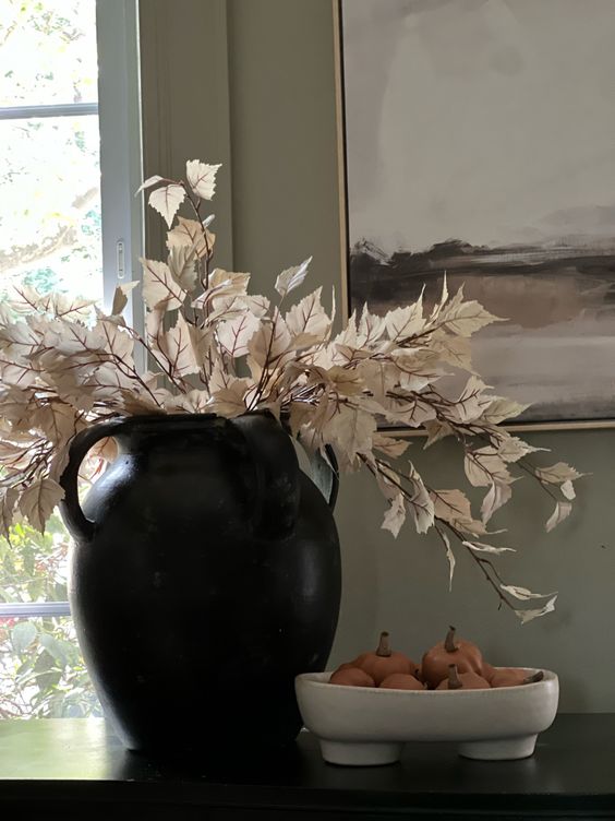 a black vase with white dried leaves, a white bowl with mini pumpkins as Thanksgiving decor