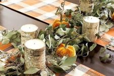 a bold fall or Thanksgiving tablescape with a eucalyptus runner and orange velvet pumpkins, candles in bark and bright plaid placemats
