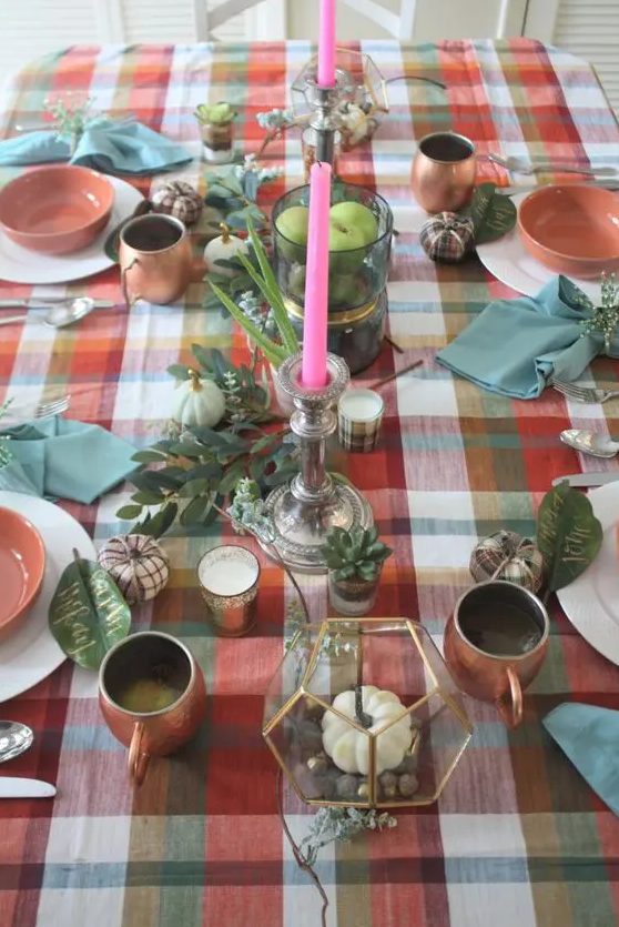 a bright Thanksgiving tablescape with a bold plaid tablecloth, greenery, pumpkins, pink candles, copper mugs, white and pink porcelain