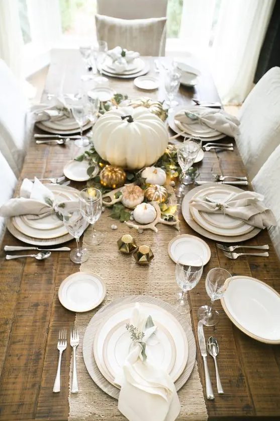 a chic Thanksgiving table with a burlap runner, white porcelain, white pumpkins, antlers, lights and greenery looks stylish