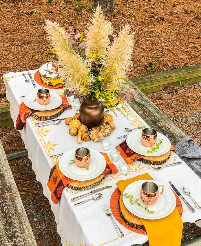 a cool forest Thanksgiving tablescape with orange placemats, wood slices, bold napkins, a grass and foliage centerpiece and copper mugs