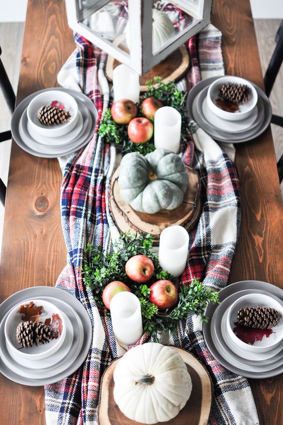a cozy and lovely woodland Thanksgiving table setting with a plaid runner, apples and greenery, pillar candles and heirloom pumpkins, large pinecones