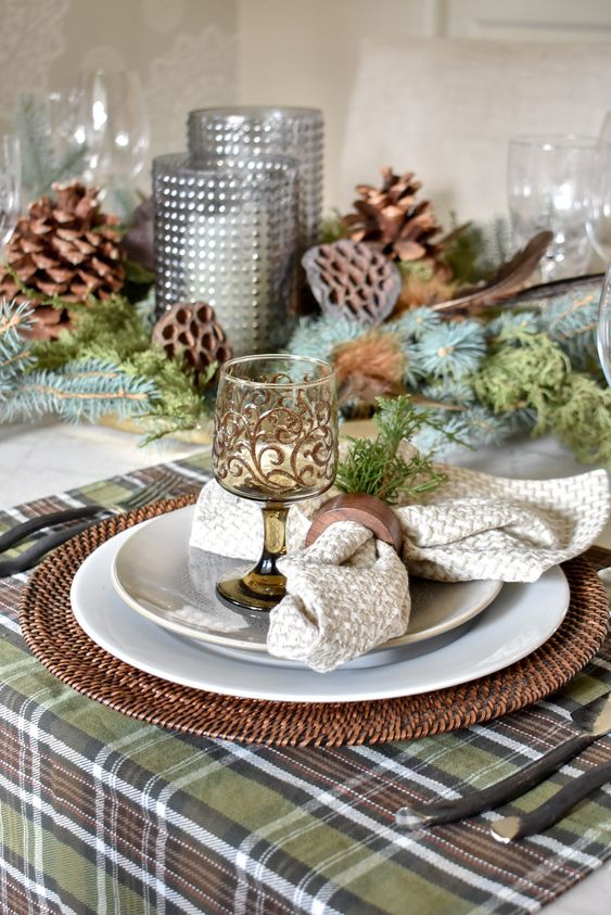 a green and brown Thanksgiving table setting with plaid and neutral linens, patterned glasses, pinecones and lotus, candles and evergreens