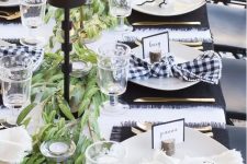 a modern black and white Thanksgiving tablescape with a greenery runner, candleholders and placemats in black, buffalo check napkins, elegant glasses and gold cutlery