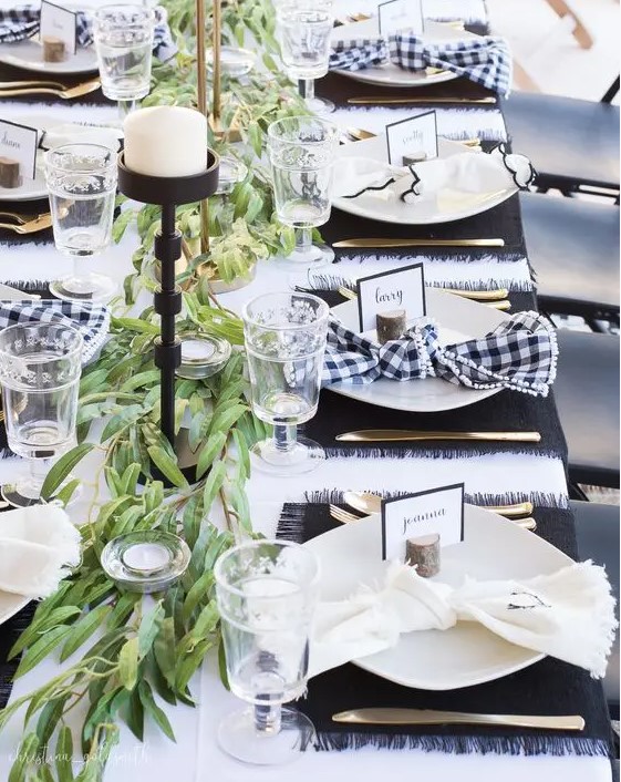 a modern black and white Thanksgiving tablescape with a greenery runner, candleholders and placemats in black, buffalo check napkins, elegant glasses and gold cutlery