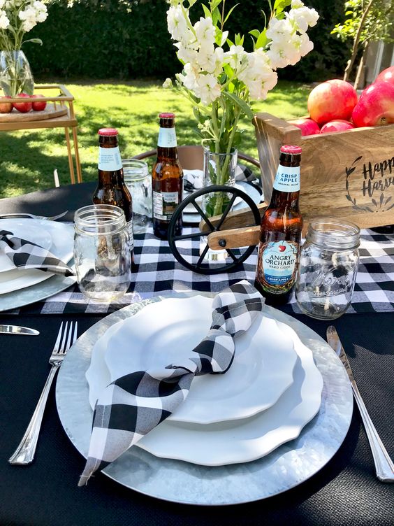 a pretty Thanksgiving table setting with a black tablecloth, buffalo check linens, a cart with apples as a centerpiece and white blooms