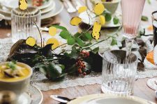 a refined woodland Thanksgiving tablescape with a lace runner, greenery, berries and gold foliage, chic neutral porcelain and ribbed glasses