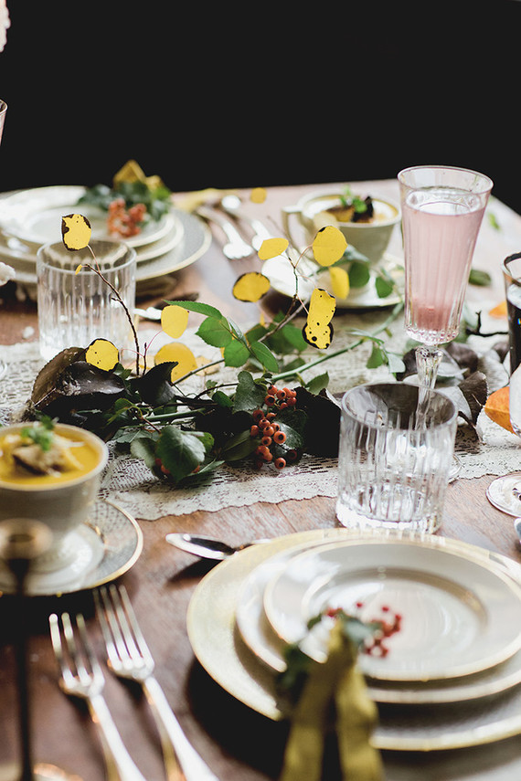 a refined woodland Thanksgiving tablescape with a lace runner, greenery, berries and gold foliage, chic neutral porcelain and ribbed glasses