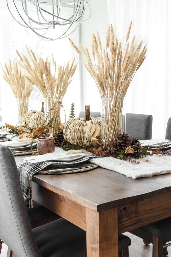 a rustic Thanksgiving tablescape with wheat in vases, pinecones and woven pumpkins, plaid napkins and a knit white runner