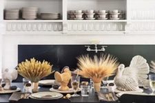 a stylish modern Thanksgiving tablescape with taupe napkins and table runners, wheat in vases, turkeys, candles and gold cutlery
