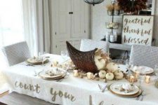 a white Thanksgiving tablescape with neutral linens, a woven cornucopia with white pumpkins and gilded nuts, gold chargers and cutlery