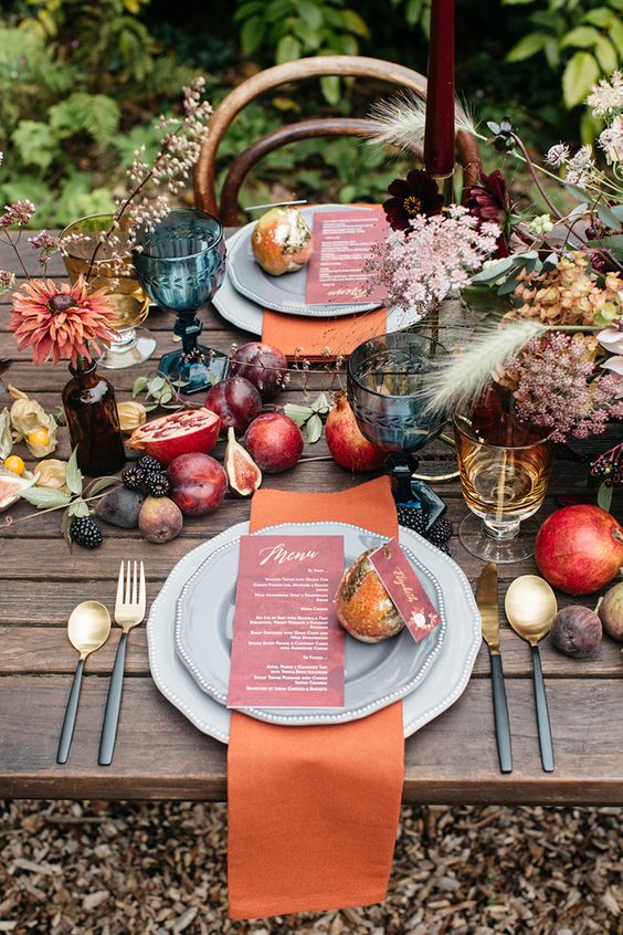 a woodland lux Thanksgiving table setting with rust napkins, fresh fruit on the table, neutral blooms and grasses plus colored glasses