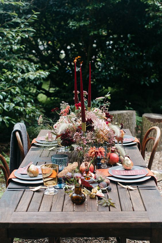 a woodland lux Thanksgiving tablescape with a super lush floral centerpiece, tall and thin burgundy candles, fresh fruit on the table and orange napkins