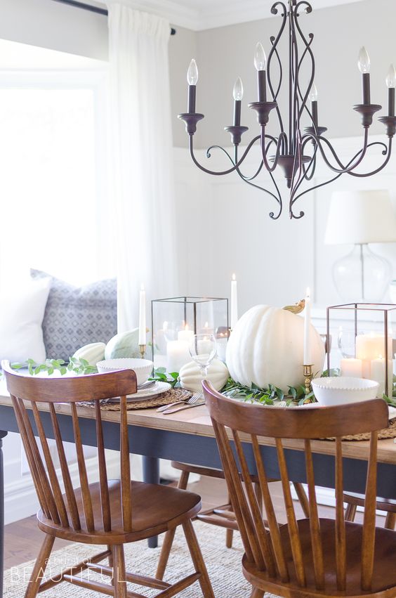 lovely white Thanksgiving tablescape with large candle lanterns, large white pumpkins, white porcelain, woven chargers and some greenery