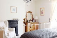 04 a chic vintage bedroom with a metal built-in fireplac,e a white metal bed, a stained dresser, a vintage chair and a beautiful crystal chandelier