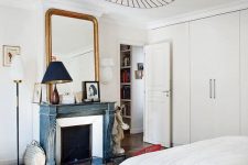 06 a serene Parisian bedroom with built-in wardrobes, a fireplace with a blue mantel, a large mirror in a gilded frame and a bed with neutral bedding