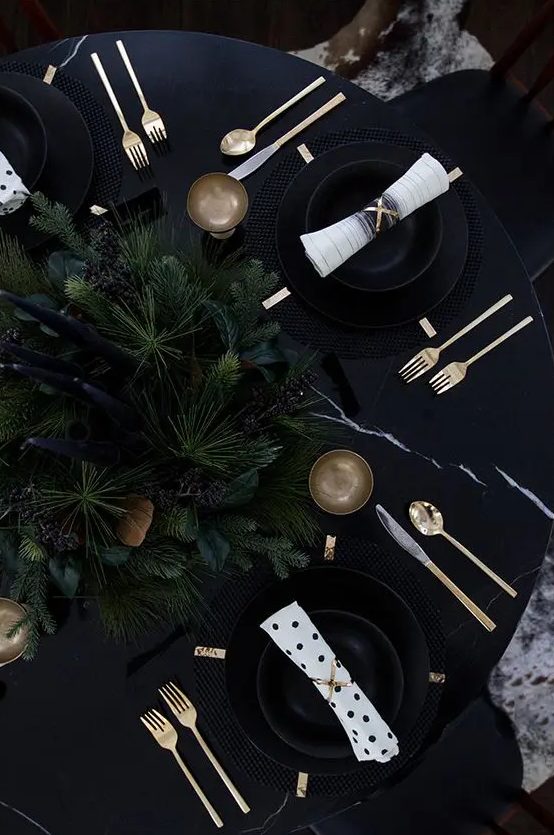 a moody and very elegant Christmas tablescape with an evergreen centerpiece, tall and thin candles, gold cutlery and candlholders, black plates and chargers