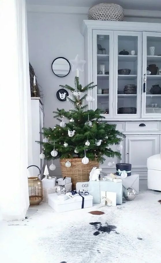 a small Nordic Christmas tree with lights and white and silver ornaments placed in a basjet and with neutral gift boxes under it is a chic idea for a modern space