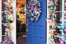 a Christmas entrance with a bright ornament garland, lights, evergreens, a candle lantern with bright decor is a cool idea