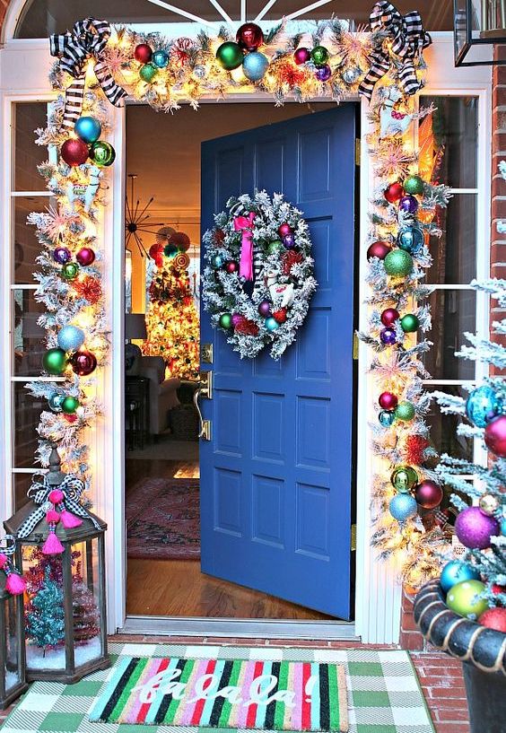 a Christmas entrance with a bright ornament garland, lights, evergreens, a candle lantern with bright decor is a cool idea