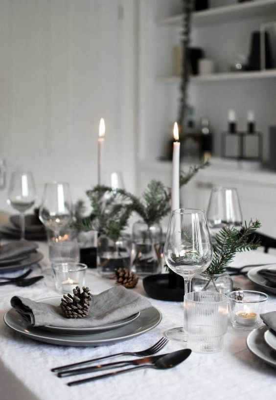 a Scandinavian Christmas tablescape with tall and thin candles in black candleholders, grey plates and napkins, pinecones and evergreens